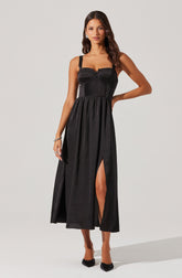 A woman is wearing a long, sleeveless black dress with wide straps. The bodice of the dress features a textured, ribbed pattern with a sweetheart neckline. The waist is fitted, and the skirt flows down to the ankles with a high slit on one side, revealing her leg. She is also wearing black pointed-toe high heels.